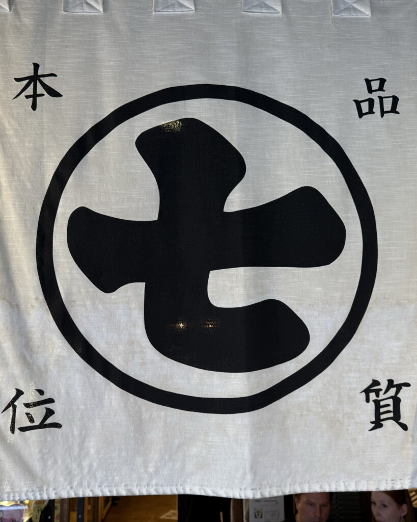 Noren (the signage fabric that hangs in front of doors in shops in Japan) for Nanaya. The noren is white background, with the Japanese kanji for number 7 (nana means 7) in black in a black circle. There are four kanji in each corner.
