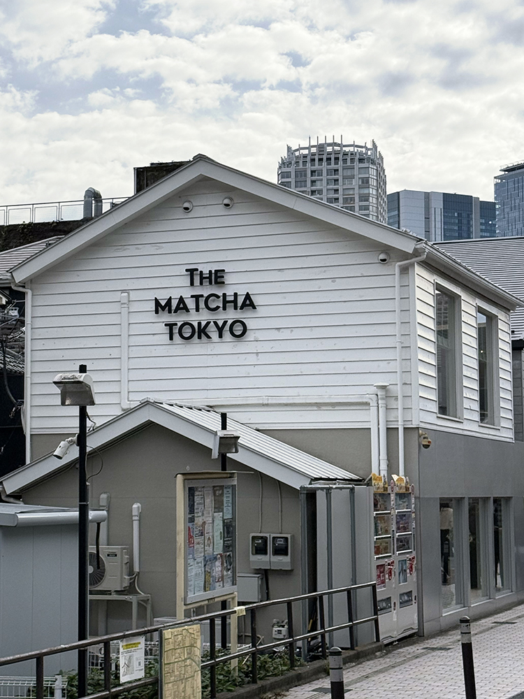 Photo of the builidng of The Matcha Tokyo in Omotesando. It's a big building, white board and windows on the second floor. Grey paint/cement on ground floor. Black lettering "The Matcha Tokyo" on the side facing Omotesando-dori