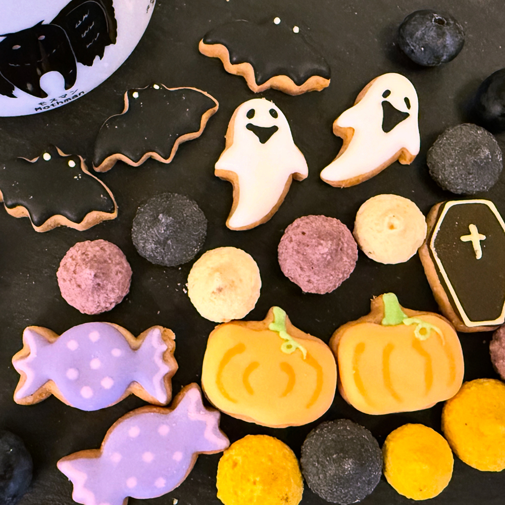 Flat lay of decorative Halloween-themed cookies from Dean & DeLuca in Tokyo, including pumpkins, ghosts, bats, coffins, wrapped treats, and mini macarons. Cozy autumn dessert display in warm orange and black tones, with a Mothman-themed cup in the background.