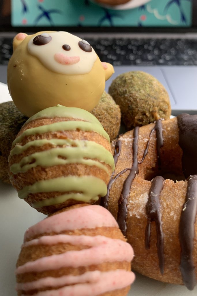 Donut assortment from Floresta donuts with a traditional chocolate drizzle, mitarashi dango-style (3 donut holes on a skewer). One is decorated like a animal face. A latte with a marshmallow shaped pig in the background.