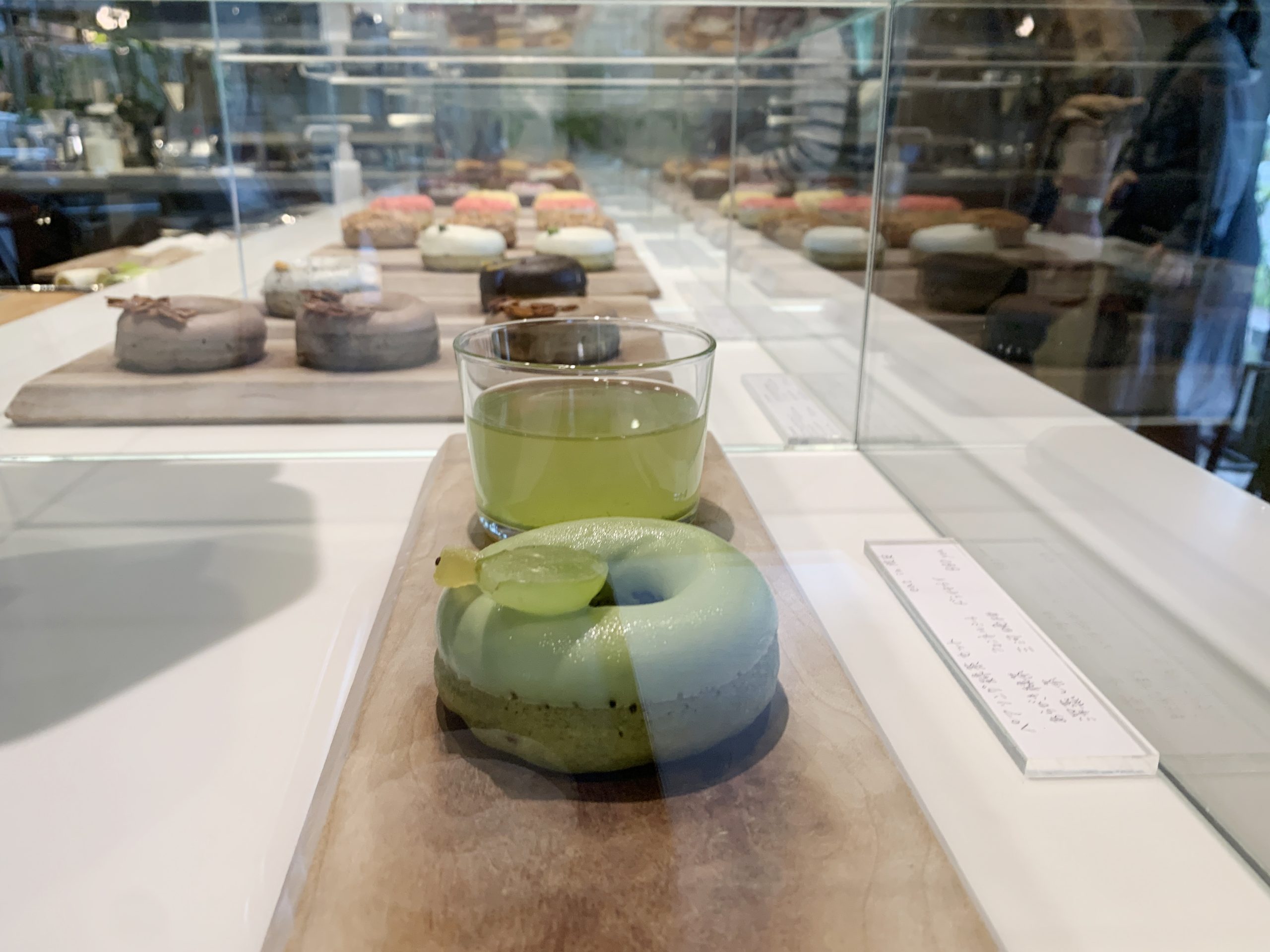 A lime donut and a glass cold green tea inside a display case at Hocus Pocus Donuts. In the case behind, more donuts are displayed like pastry jewels.